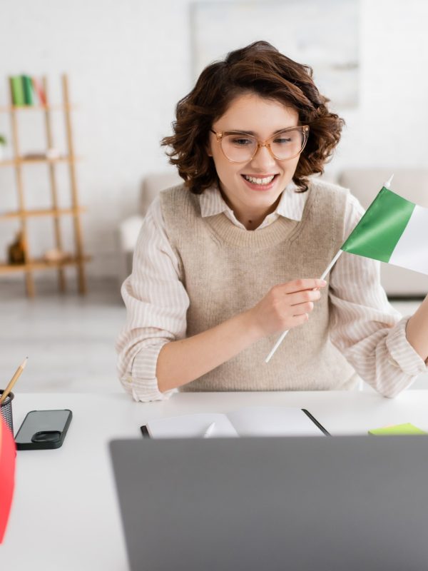 young teacher holding flag of Italy near laptop and textbook of French language while working at
